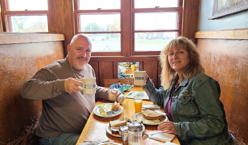 A man and woman sit at a wooden table, smiling and holding mugs, with breakfast items in front of them.