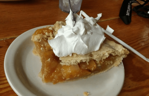 A slice of apple pie topped with whipped cream on a white plate, resting on a wooden table.