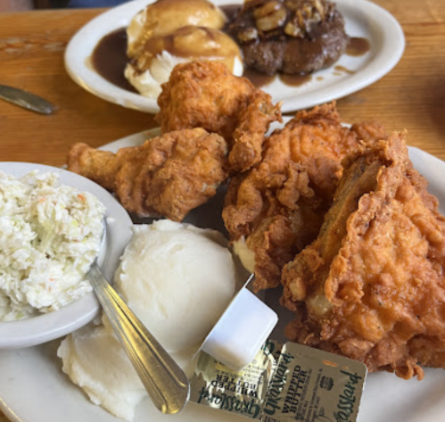 A plate of fried chicken with mashed potatoes, coleslaw, and a butter packet, with desserts in the background.