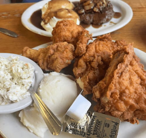 A plate of fried chicken with mashed potatoes, coleslaw, and a butter packet, with desserts in the background.