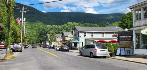 A quaint street lined with shops and cars, set against a backdrop of green mountains and a blue sky.