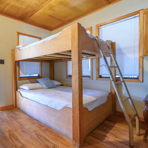 A wooden bunk bed in a room with two mattresses, a ladder, and windows with blinds.