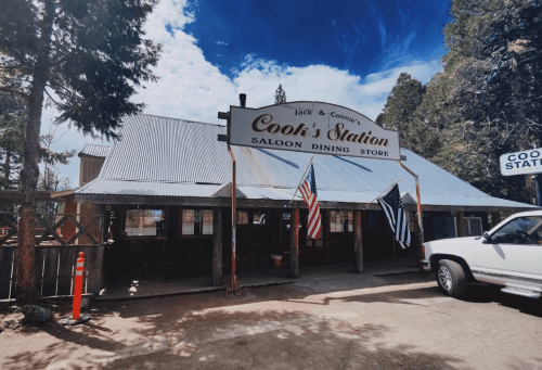 A rustic building with a metal roof, featuring a sign for Cook's Station and American flags outside.