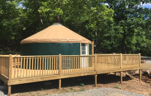 A green and tan yurt on a wooden deck, surrounded by trees and gravel, under a clear blue sky.