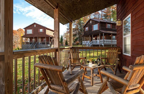 A cozy porch with wooden chairs overlooks two rustic cabins surrounded by autumn foliage.