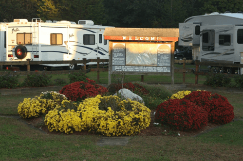 A flower garden with yellow and red blooms in front of a welcome sign and parked RVs in a campground.
