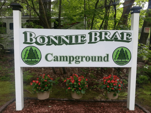 Sign for Bonnie Brae Campground, featuring green text and decorative trees, with flower pots below.
