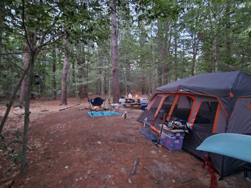 A campsite in a wooded area featuring a tent, chairs, a picnic table, and a small fire.