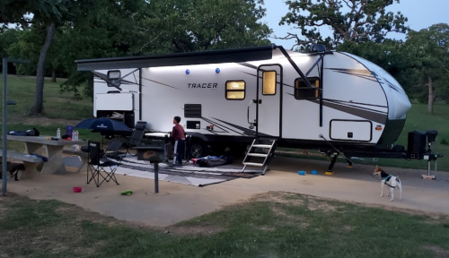 A camper trailer parked in a grassy area, with a child playing outside and a dog nearby. Evening light casts a warm glow.