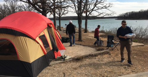 A campsite by a lake with a red tent, people setting up, and trees in the background under a cloudy sky.