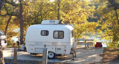 A small white Scamp trailer parked near a lake, surrounded by trees with autumn foliage.