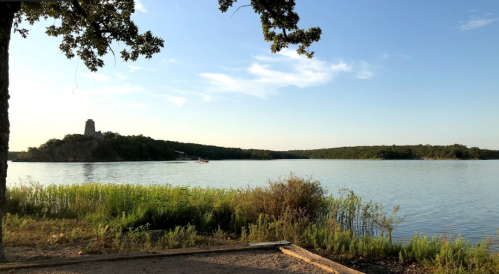 A serene lake scene with lush greenery, a distant tower on a hill, and a clear blue sky.