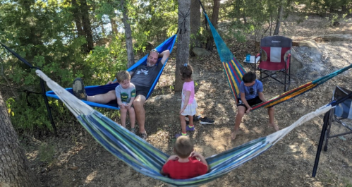 A family enjoys a relaxing day outdoors in hammocks by a lake, with children playing and adults lounging.