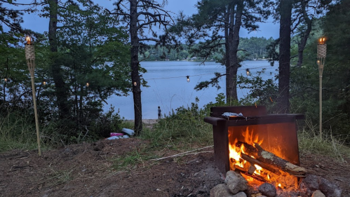 A cozy campfire with logs burning, surrounded by trees, overlooking a calm lake at dusk. Tiki torches are lit nearby.