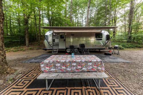 A silver Airstream trailer parked in a wooded area, with a picnic table and outdoor seating set up nearby.