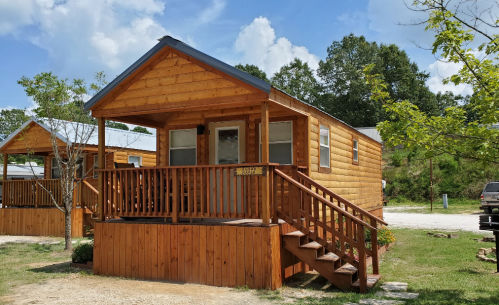 A wooden cabin with a porch, surrounded by greenery and blue skies, in a serene outdoor setting.