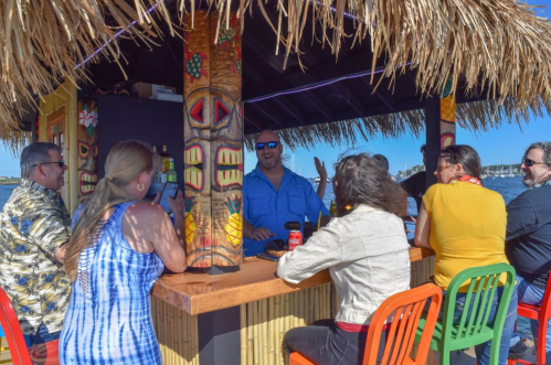 A group of people enjoying drinks at a tiki bar by the water, with colorful decor and a thatched roof.