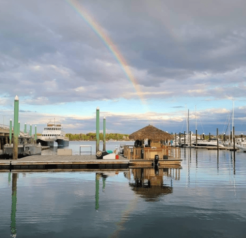 A serene marina scene with a rainbow, boats, and a thatched-roof hut reflected in calm waters.