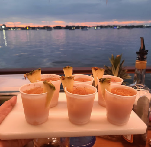 A hand holds a tray of seven colorful shot glasses with pineapple garnishes, set against a sunset over water.