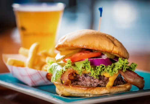 A juicy burger with lettuce, tomato, onion, bacon, and cheese, served with fries and a glass of beer in the background.