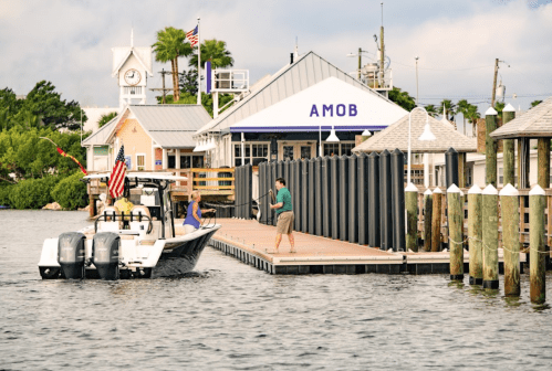 A boat approaches a dock with a sign reading "AMOB," while a person fishes nearby. Flags wave in the background.