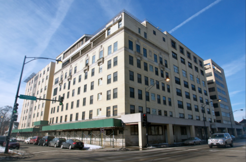 A large, multi-story building with a beige facade, scaffolding, and clear blue skies in the background.