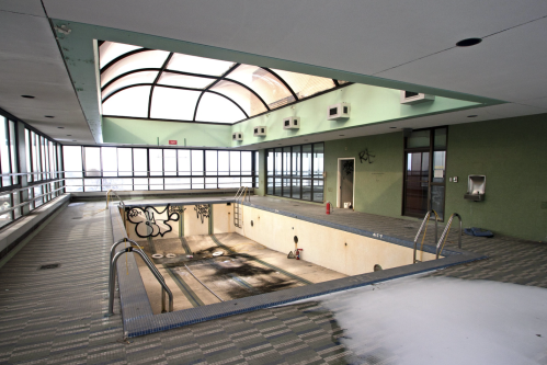 Abandoned indoor pool with graffiti, empty water basin, and large windows letting in natural light.