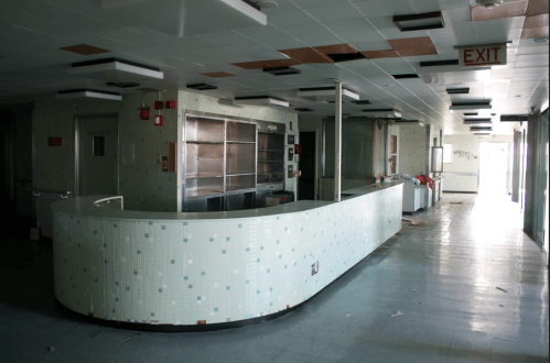 Abandoned interior of a hospital reception area with a curved counter and scattered debris. Exit sign visible.