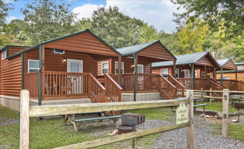Three wooden cabins with porches, surrounded by trees and gravel, under a partly cloudy sky.