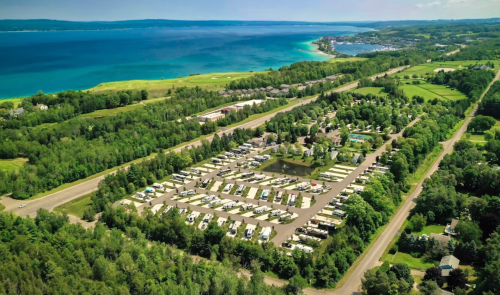 Aerial view of a lush campground near a blue lake, with rows of RVs and green landscapes under a clear sky.
