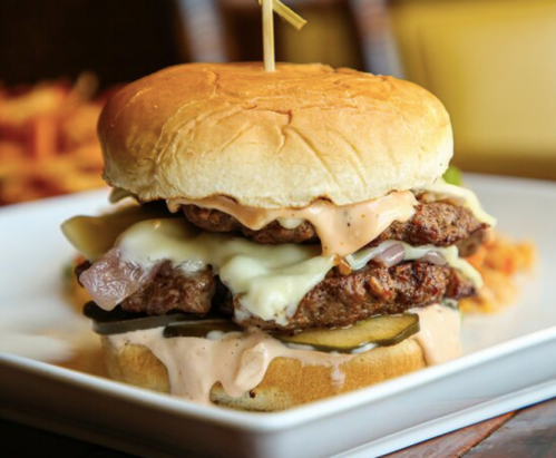 A stacked burger with two patties, melted cheese, pickles, onions, and sauce, served on a soft bun.