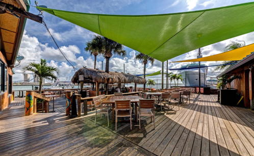 Outdoor dining area with wooden tables, palm trees, and colorful shade sails against a cloudy sky.
