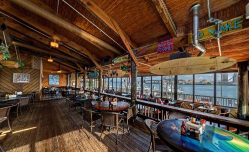 Cozy restaurant interior with wooden decor, tables set for dining, and views of a marina through large windows.