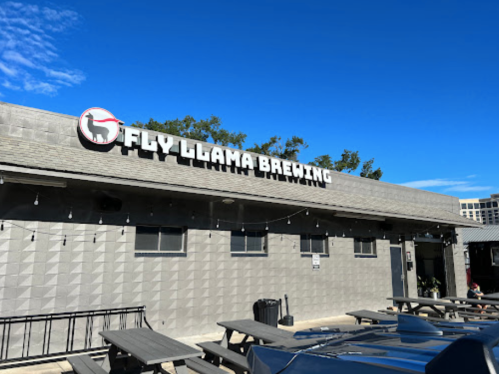 Exterior of Fly Llama Brewing with a clear blue sky, featuring picnic tables and a modern building design.