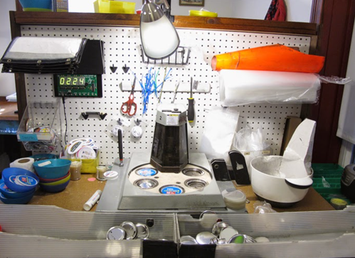 A cluttered workspace with tools, containers, and a coffee maker on a pegboard wall, featuring various supplies and equipment.