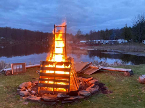 A large bonfire burns brightly in a stone circle by a lake, with trees and a campsite visible in the background.