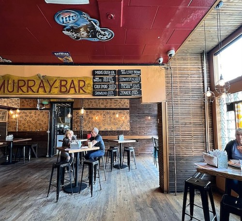Interior of a bar with wooden decor, tables, and a menu board. Two patrons are seated at a table.