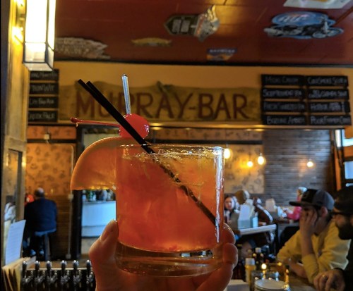A hand holds a colorful cocktail with a cherry and orange slice, set against a bar backdrop with patrons in the background.