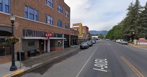 Street view of a small town featuring brick buildings, a bar sign, parked cars, and trees lining the road.