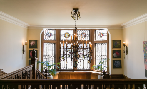 A grand staircase with a chandelier, stained glass windows, and artwork on the walls.