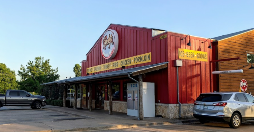 A red and wooden building with signs for BBQ, beer, and goods, featuring a parking lot with two vehicles.
