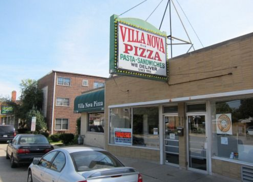 Exterior of Villa Nova Pizza restaurant with a large sign, parked cars, and a nearby building.