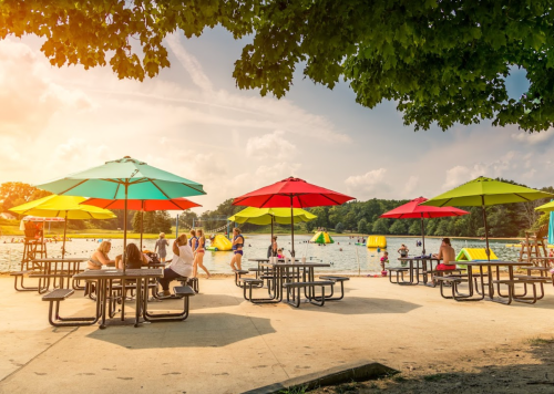 People relax under colorful umbrellas by a lake, with inflatable attractions and a sunny sky in the background.
