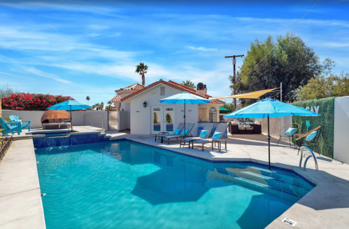 A sunny pool area with blue umbrellas, lounge chairs, and a hot tub surrounded by palm trees and vibrant flowers.