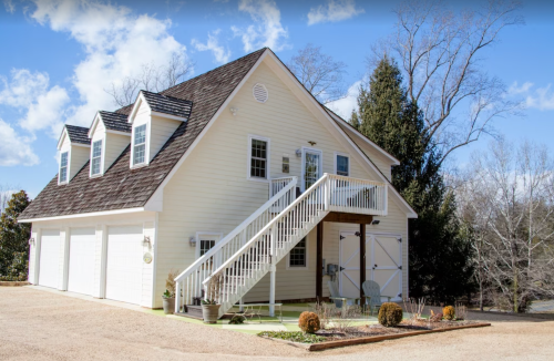 A two-story white house with a staircase leading to the entrance, surrounded by a gravel driveway and trees.