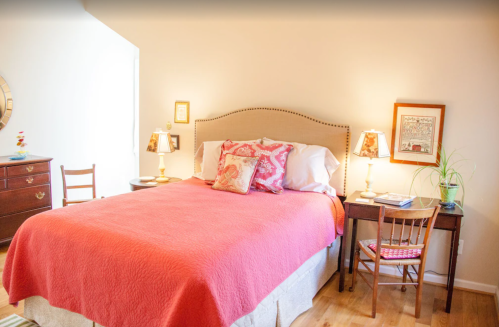 Cozy bedroom with a pink bedspread, bedside lamps, a small desk, and a wooden dresser. Warm, inviting atmosphere.