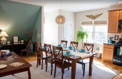 A bright dining area with a wooden table set for dinner, a desk in the corner, and a large window with plants.