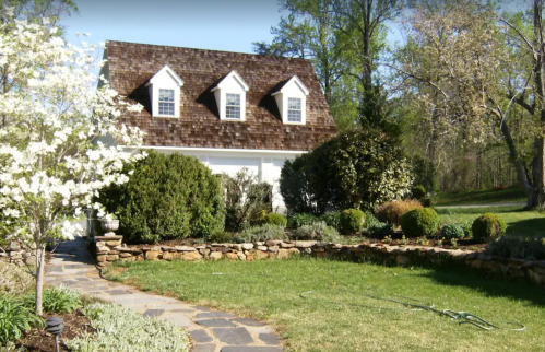 A charming house with a sloped roof, surrounded by lush greenery and flowering plants in a sunny garden.