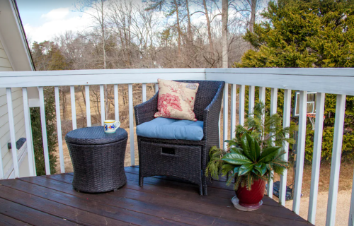 A cozy balcony with a wicker chair, blue cushion, small table, and a potted plant, surrounded by trees.