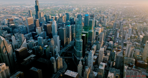 Aerial view of a bustling city skyline with tall buildings and green spaces, bathed in warm sunlight.